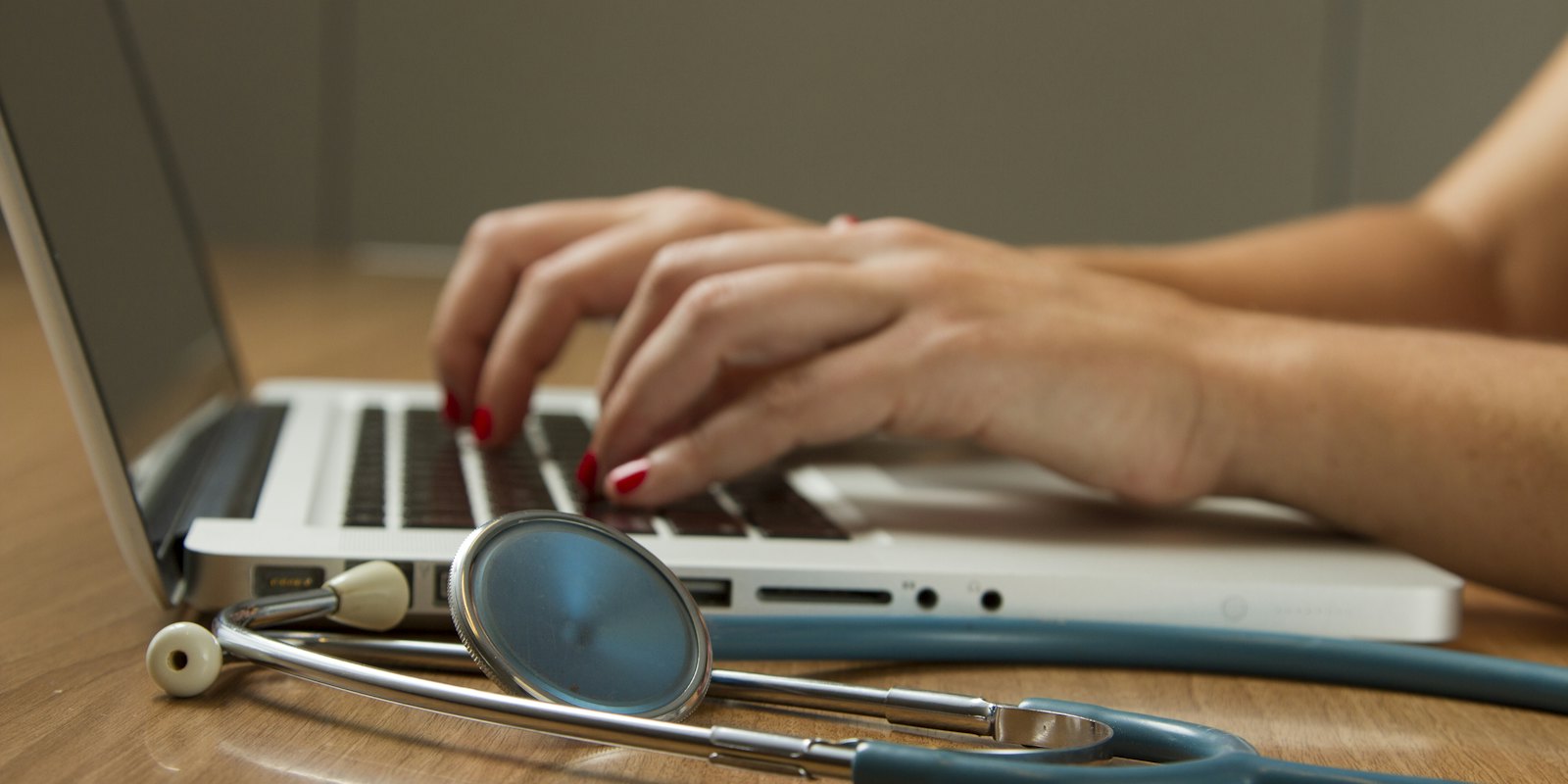 Hands typing on a laptop with a stethoscope on the desk